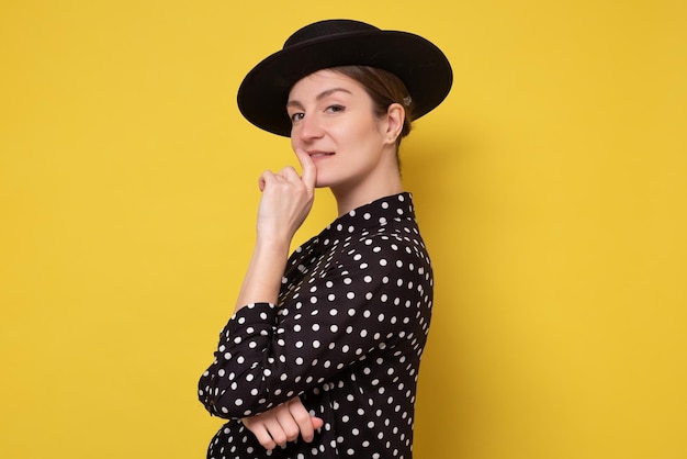 Stijlvolle blanke vrouw met een zwarte hoed die met vertrouwen naar de camera kijkt. Studio opname op gele muur.