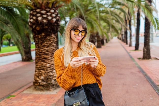 Stijlvolle blonde vrouw geniet van weekend in Spanje, met behulp van smartphone