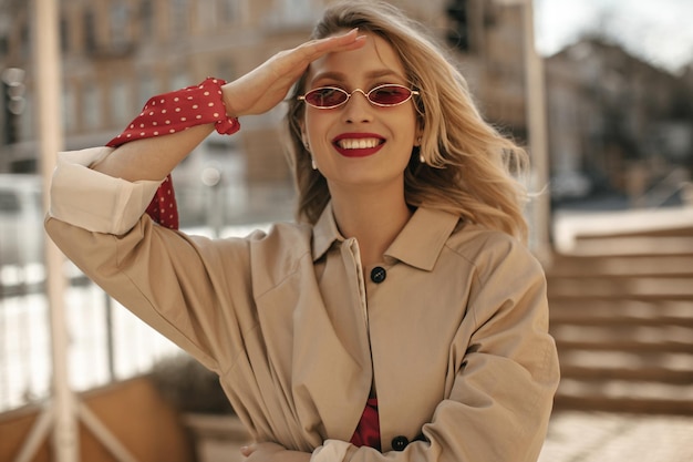 Stijlvolle blonde vrouw in beige trenchcoat en rode zonnebril glimlacht oprecht portret van mooie dame met heldere lippen buitenshuis