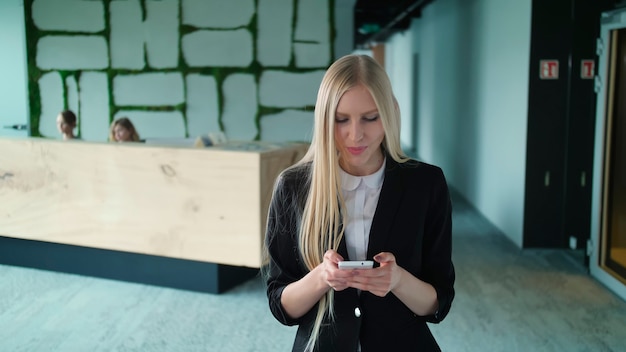 Stijlvolle blonde vrouw in jas die in de hedendaagse kantoorzaal staat en smartphone surft met een glimlach