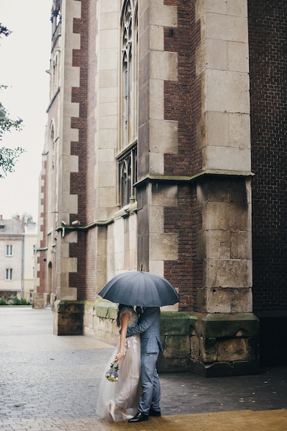 Stijlvolle bruid en bruidegom zoenen onder de paraplu op de achtergrond van de oude kerk in de regen provence bruiloft