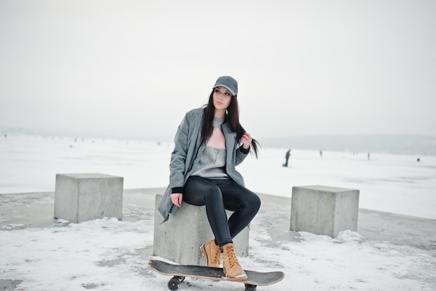 Stijlvolle brunette meisje in grijze pet casual street style met skate board op winterdag