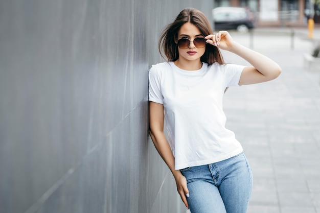 Stijlvolle brunette meisje met witte t-shirt en bril poseren tegen straat stedelijke kledingstijl