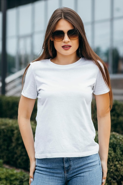 Stijlvolle brunette meisje met witte t-shirt en bril poseren tegen straat stedelijke kledingstijl