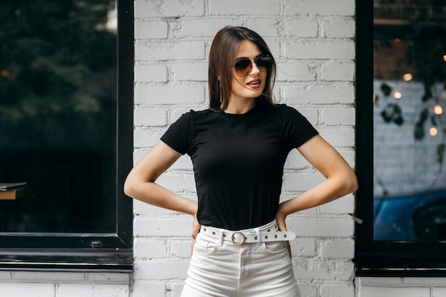 Stijlvolle brunette meisje met zwarte t-shirt en bril poseren tegen straat stedelijke kledingstijl