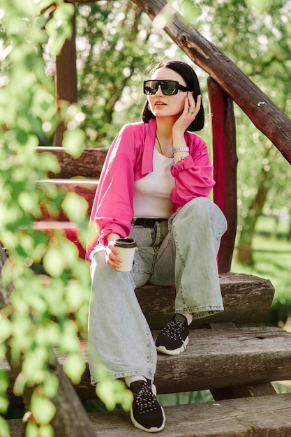 Stijlvolle brunette vrouw in zonnebrillen en casual kleding met een kopje koffie en buiten in het park zitten
