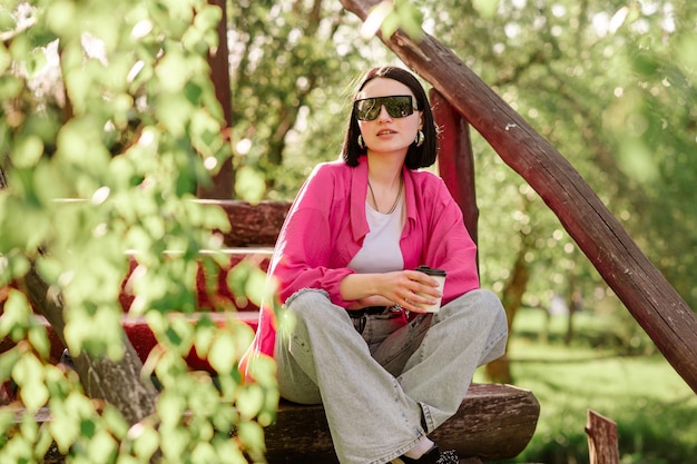 Stijlvolle brunette vrouw in zonnebrillen en casual kleding met een kopje koffie en buiten in het park zitten