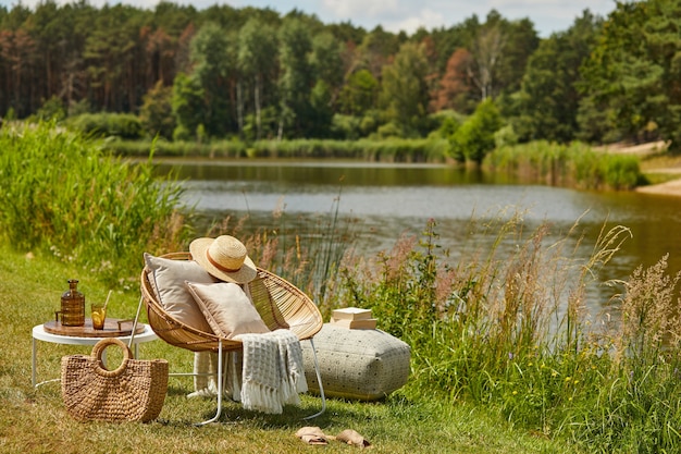 Foto stijlvolle compositie van buitentuin aan het meer met design rotan fauteuil, salontafel, plaid, kussens, drankjes en elegante accessoires.