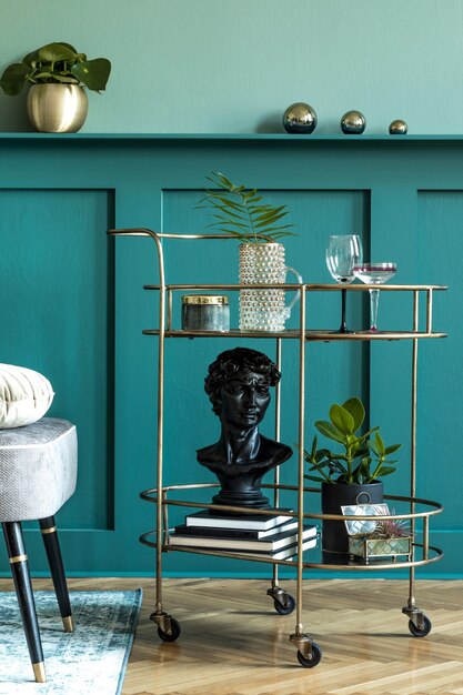 Stijlvolle en moderne compositie van woonkamer met design gouden drankkast, planten, boeken en elegante persoonlijke accessoires. Groene wandbekleding met plank. Moderne woondecoratie. Sjabloon.