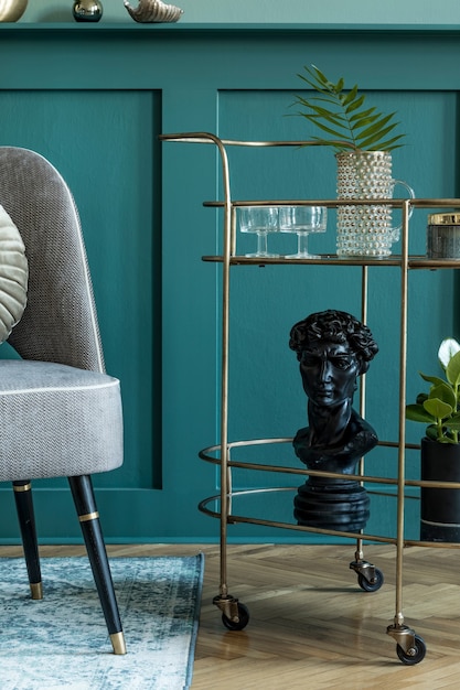 Stijlvolle en moderne compositie van woonkamer met grijze design fauteuil, gouden drankkast, planten en elegante persoonlijke accessoires. Grijze wandbekleding met plank. Moderne woondecoratie. Sjabloon.