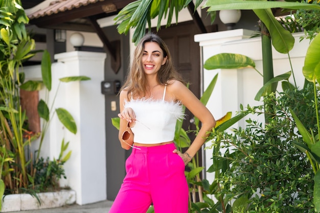 Foto stijlvolle, gebruinde, mooie vrouw in een zonnebril, roze broek en witte top die buiten poseert