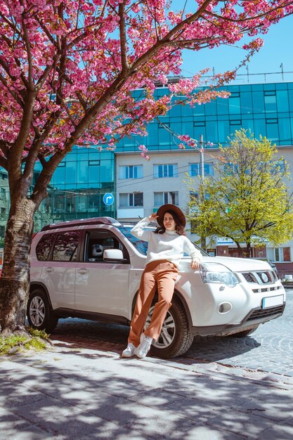 Stijlvolle glimlachende mooie vrouw die in de buurt van een bloeiende sakura-boom in de auto staat op de achtergrond, de lente komt eraan