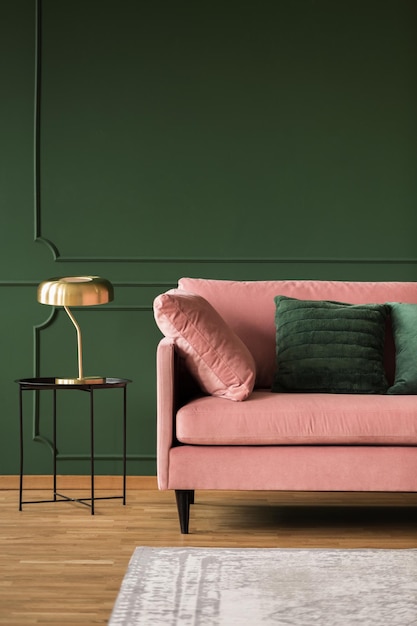 Stijlvolle gouden lamp op industriële tafel naast roze bank met smaragdgroene kussens in donkergroene woonkamer interieur kopie ruimte op lege muur