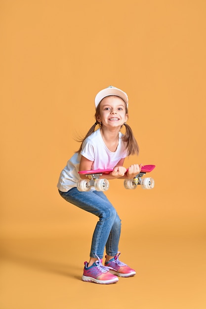 Stijlvolle grappig meisje draagt een wit t-shirt, blauwe spijkerbroek en sneakers, met skateboard over gele muur