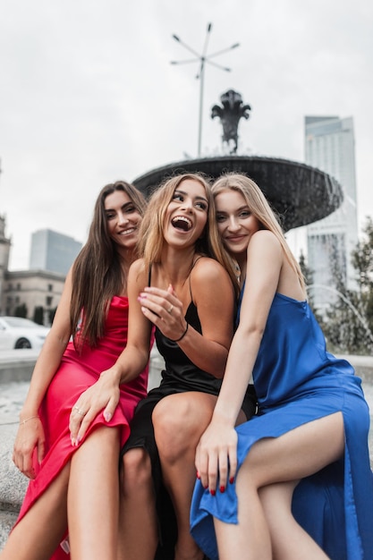 Stijlvolle grappige mooie modieuze vrouwelijke vriendinnen met sexy benen in modejurken zitten bij de fontein in de stad