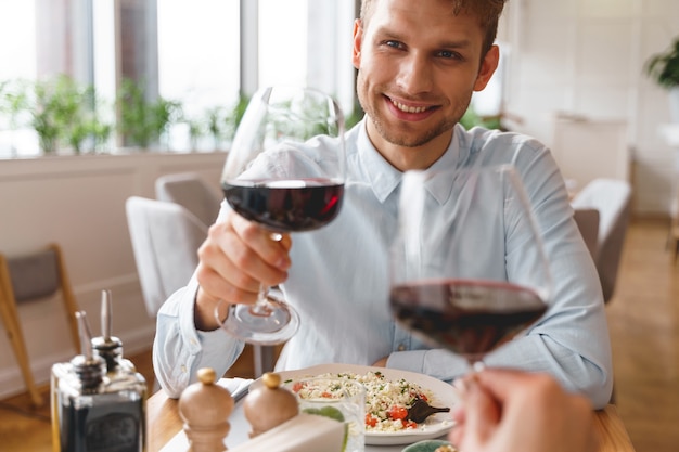 Stijlvolle heer die een glas alcoholische drank vasthoudt en naar de dame met een glimlach kijkt terwijl hij aan tafel zit met heerlijk eten