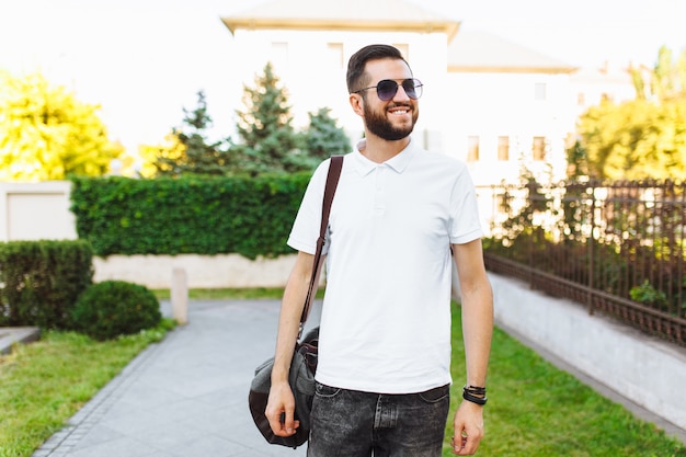 Stijlvolle hipster met een baard in een wit t-shirt, met een reistas in zijn handen die door de stad lopen