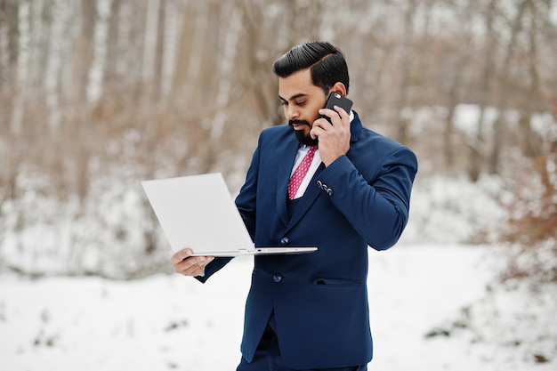 Stijlvolle Indiase baard zakenman in pak geposeerd op winterdag buiten met laptop in handen sprekend op mobiele telefoon