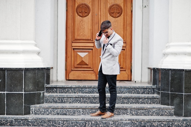 Stijlvolle Indiase man met bindi op voorhoofd en bril draagt een grijs pak dat buiten tegen de deur van het gebouw staat en op mobiele telefoon spreekt