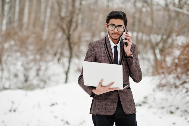 Stijlvolle indiase student man in pak en bril poseerde op winterdag buiten met laptop in handen die aan de telefoon sprak