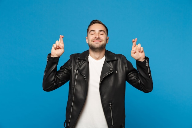 Stijlvolle jonge, bebaarde man in zwart leren jas wit t-shirt wachten op speciaal moment houden vingers gekruiste ogen gesloten geïsoleerd op blauwe muur achtergrond. Mensen oprechte emoties concept Mock up