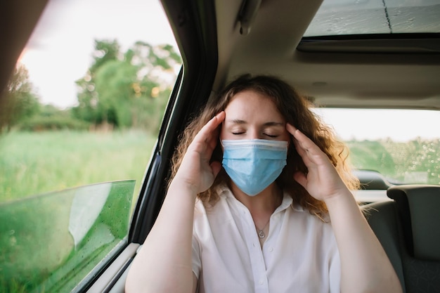 Stijlvolle jonge carly-vrouw met medisch masker heeft hoofdpijn terwijl ze op de achterbank van de auto zit