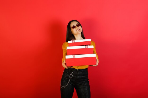 Stijlvolle jonge gelukkig lachende vrouw in rode zonnebril houdt drie rood-witte geschenkverpakkingen geïsoleerd op rood studiooppervlak.