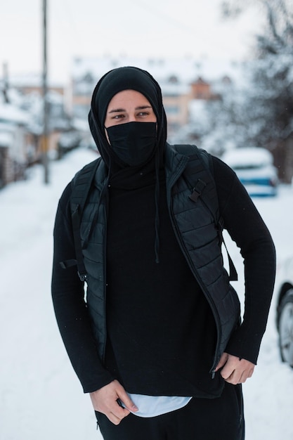 Stijlvolle jonge kerel met een zwart masker in zwarte bovenkleding met een modieus vest, hoodie, hoed en rugzak loopt op een winterdag met sneeuw