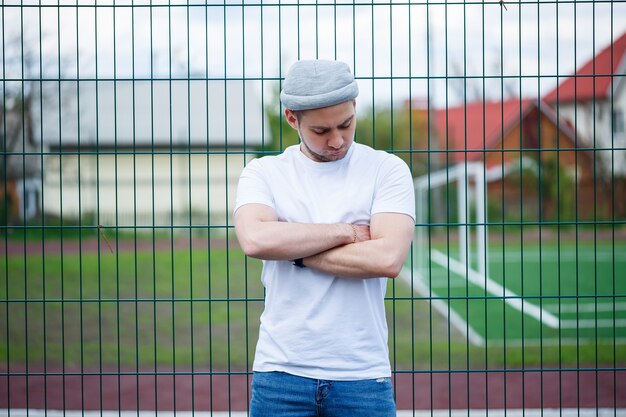 Stijlvolle jonge man, een man gekleed in een wit leeg t-shirt en een grijze hoed die tegen de achtergrond van een raster van een voetbalveld staat. Stedelijke kledingstijl, modern modieus beeld. Herenmode