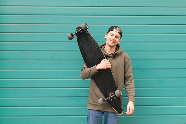 Stijlvolle jonge man in een hoodie en pet, staande met een skate in zijn handen tegen de achtergrond van een turquoise muur