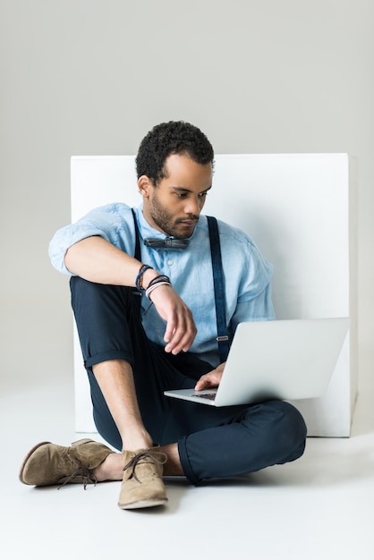 Stijlvolle jonge man zitten en met behulp van laptop op grijs