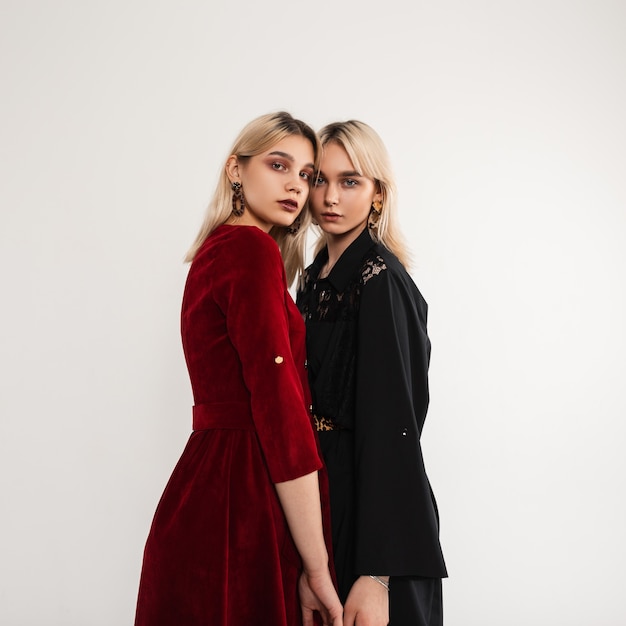 Stijlvolle jonge mooie vrouwen met blond haar in mode jurken binnenshuis. Twee modieuze vriendinnen in vintage kleding in retro stijl poseren in de buurt van muur