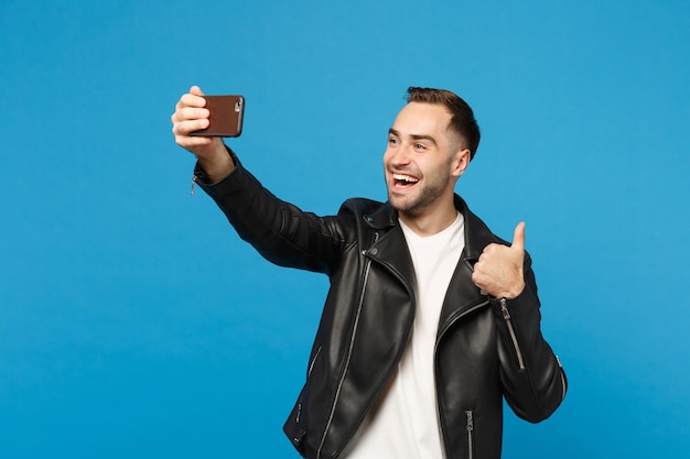 Stijlvolle jonge ongeschoren man in zwarte jas wit t-shirt oing selfie geschoten op mobiele telefoon geïsoleerd op blauwe muur achtergrond studio portret. Mensen oprechte emoties levensstijl concept Mock up kopie ruimte