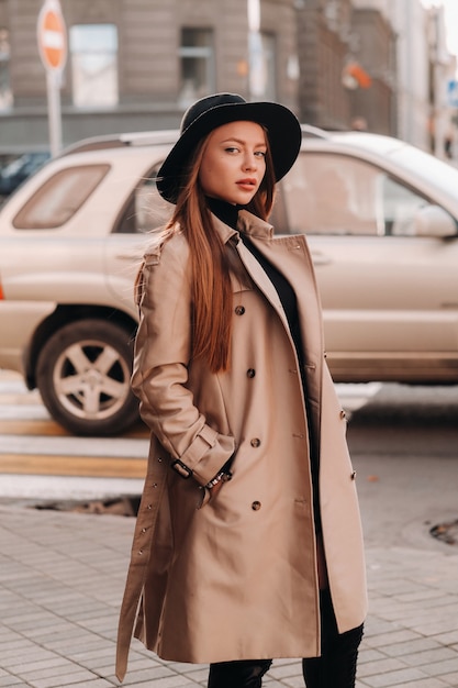Stijlvolle jonge vrouw in een beige jas in een zwarte hoed op een stadsstraat. Straatmode voor dames. Herfst kleding. Stedelijke stijl.