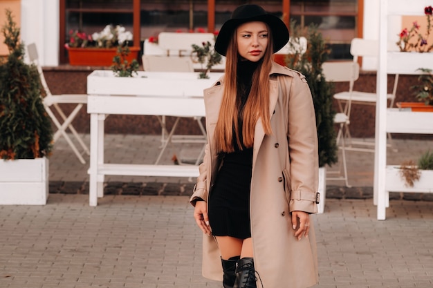 Stijlvolle jonge vrouw in een beige jas in een zwarte hoed op een straat in de stad. Street fashion voor dames. Herfstkleding. Stedelijke stijl.