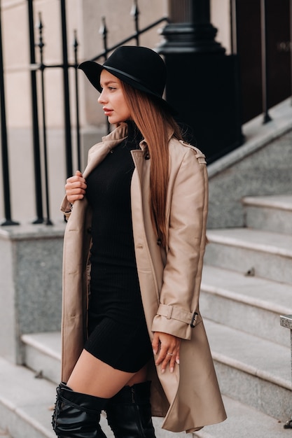 Stijlvolle jonge vrouw in een beige jas in een zwarte hoed op een straat in de stad. street fashion voor dames. herfstkleding. stedelijke stijl.