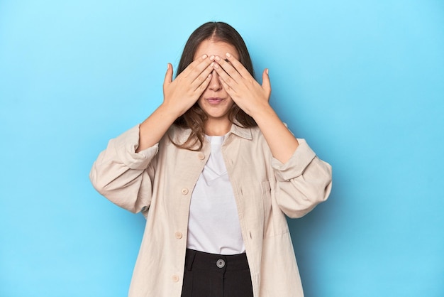 Stijlvolle jonge vrouw in een overhemd op een blauwe achtergrond bang om ogen met handen te bedekken