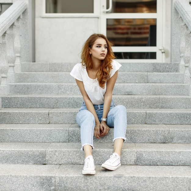 Stijlvolle jonge vrouw in een wit overhemd, spijkerbroek en sneakers zittend op de trap.