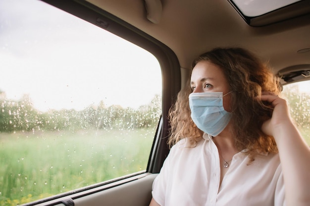 Stijlvolle jonge vrouw met een medisch masker die uit het raam kijkt terwijl ze op de achterbank van de auto zit