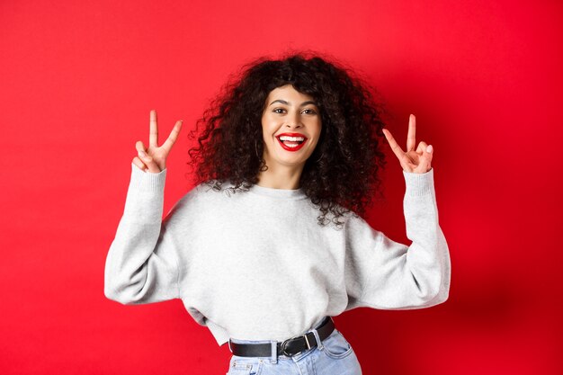 Stijlvolle jonge vrouw met krullend kapsel, gelukkig lachend en vredestekens tonend, staande in een casual sweatshirt op rode achtergrond