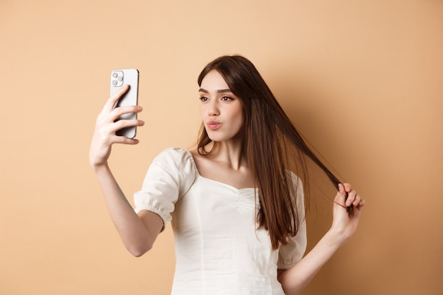 Stijlvolle jonge vrouw spelen met haar en selfie nemen op smartphone, foto maken voor sociale media, staande op beige achtergrond.