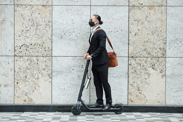 Stijlvolle jonge zakenman met paardenstaart rijden op scooter langs hoge granieten muur