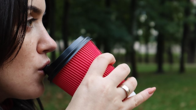 Stijlvolle jongedame drinkt 's ochtends hete koffie in rode eco-papieren beker buiten in het herfstpark. close-up zijaanzicht van jonge vrouw met afhaalmaaltijden koffiekopje.