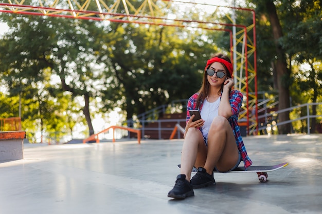 Stijlvolle jongedame gekleed in jeugd kleding zit op een skateboard en luistert naar muziek op koptelefoon op een skatepark. Zomertijd