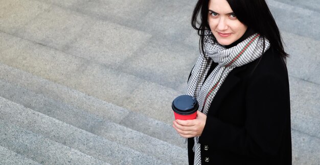 Stijlvolle jongedame in een jas en sjaal drinkt 's ochtends warme koffie in een rood eco-papieren glas buiten in een herfstpark. Portret van een jonge vrouw met een afhaalmaaltijden kopje koffie, ondiepe scherptediepte.