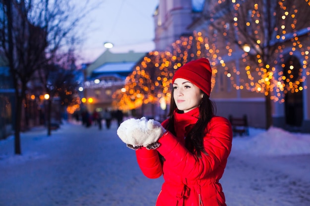 Stijlvolle jongedame in rode jas en rode gebreide muts met stapel sneeuw in haar handen. Buitenportret van een vrouw in warme, lichte kleding, stad met kerstverlichting in onscherpte op de achtergrond