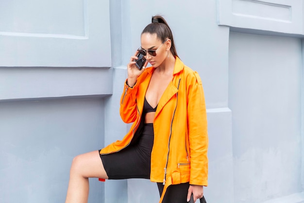 Stijlvolle jongedame op straat spreekt aan de telefoon Een mooie brunette in een fel oranje jasje en zonnebril staat tegen een grijze muur Communicatie en werken op afstand