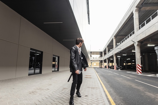 Stijlvolle jongeman in een oversized leren jas in vintage broek in coole laarzen met een modieuze zwarte rugzak loopt op straat in de buurt van de parkeerplaats. moderne coole kerel in trendy kleding buitenshuis.