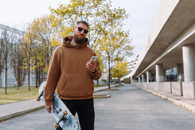 Stijlvolle jongeman met baard houdt een skateboard en smartphone vast Stedelijke plaats Ruimte kopiëren Beste kwaliteit foto