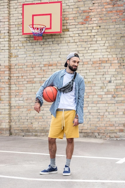 Stijlvolle jongeman met basketbalbal die op straat staat en wegkijkt